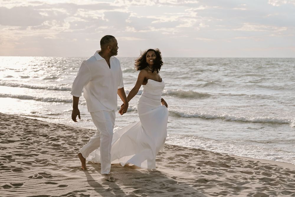 A couple in white outfits walks together along a scenic beach at sunset, creating romantic silhouettes against the ocean.