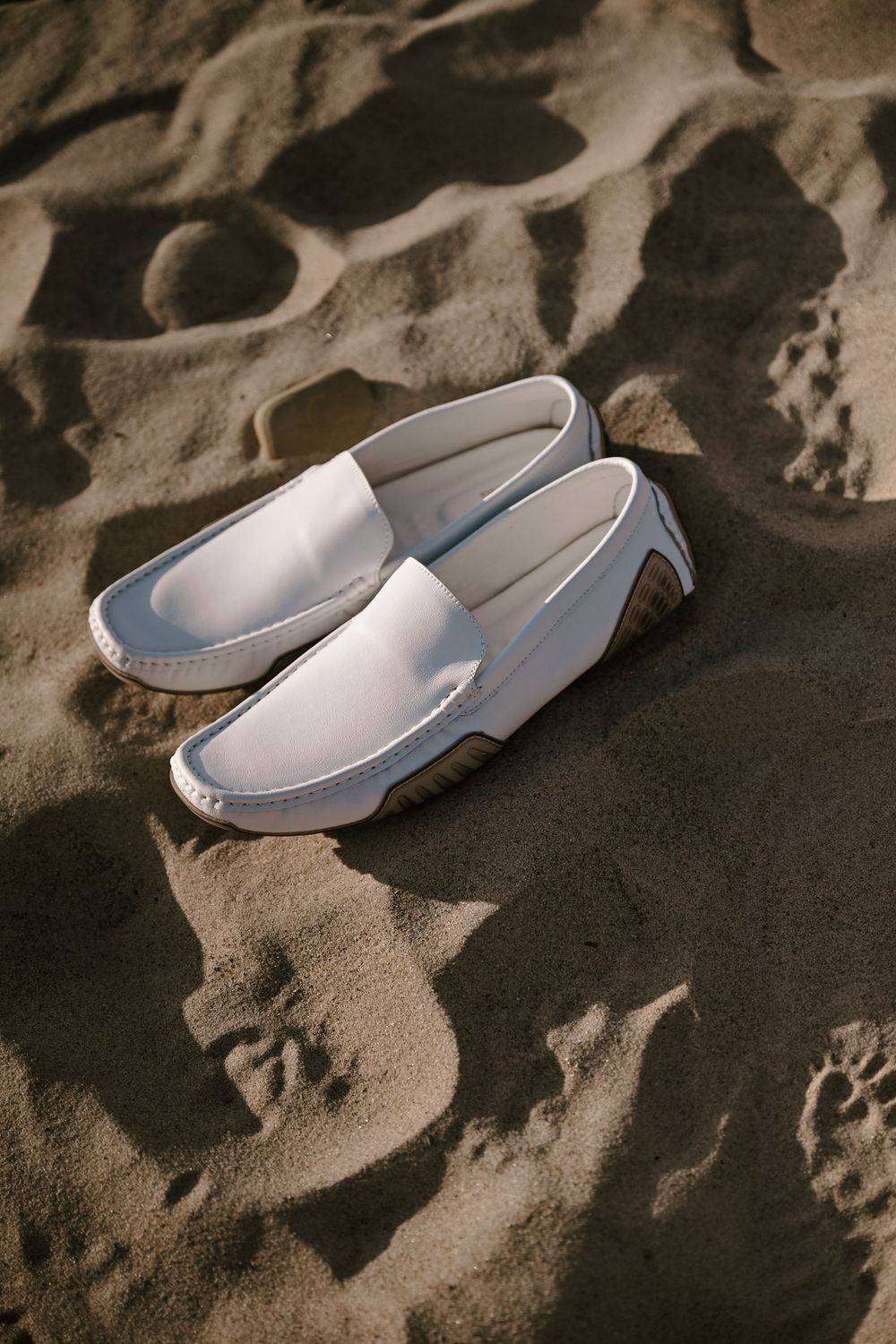 White loafers sitting on textured sand at the beach creating artistic shadows and patterns.