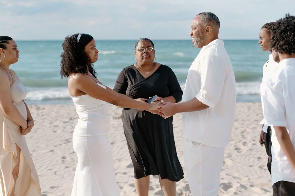 A group of people in white and black attire walking along a sunny beach with turquoise ocean waters in the background.