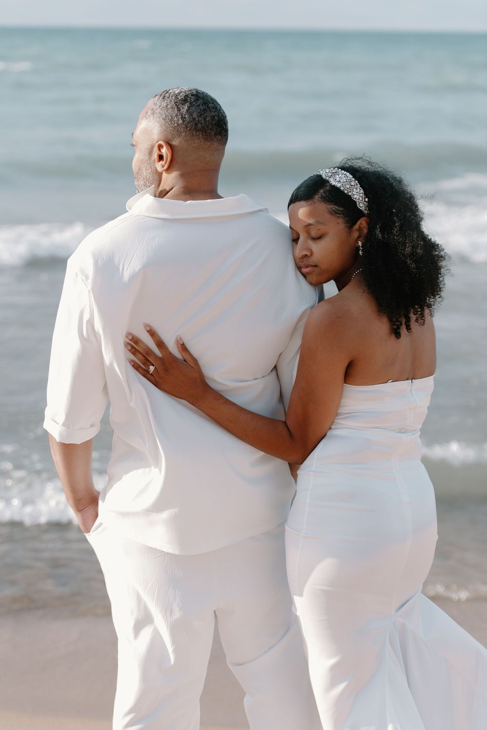 A couple in white attire embraces while walking along a beach with ocean waves in the background.