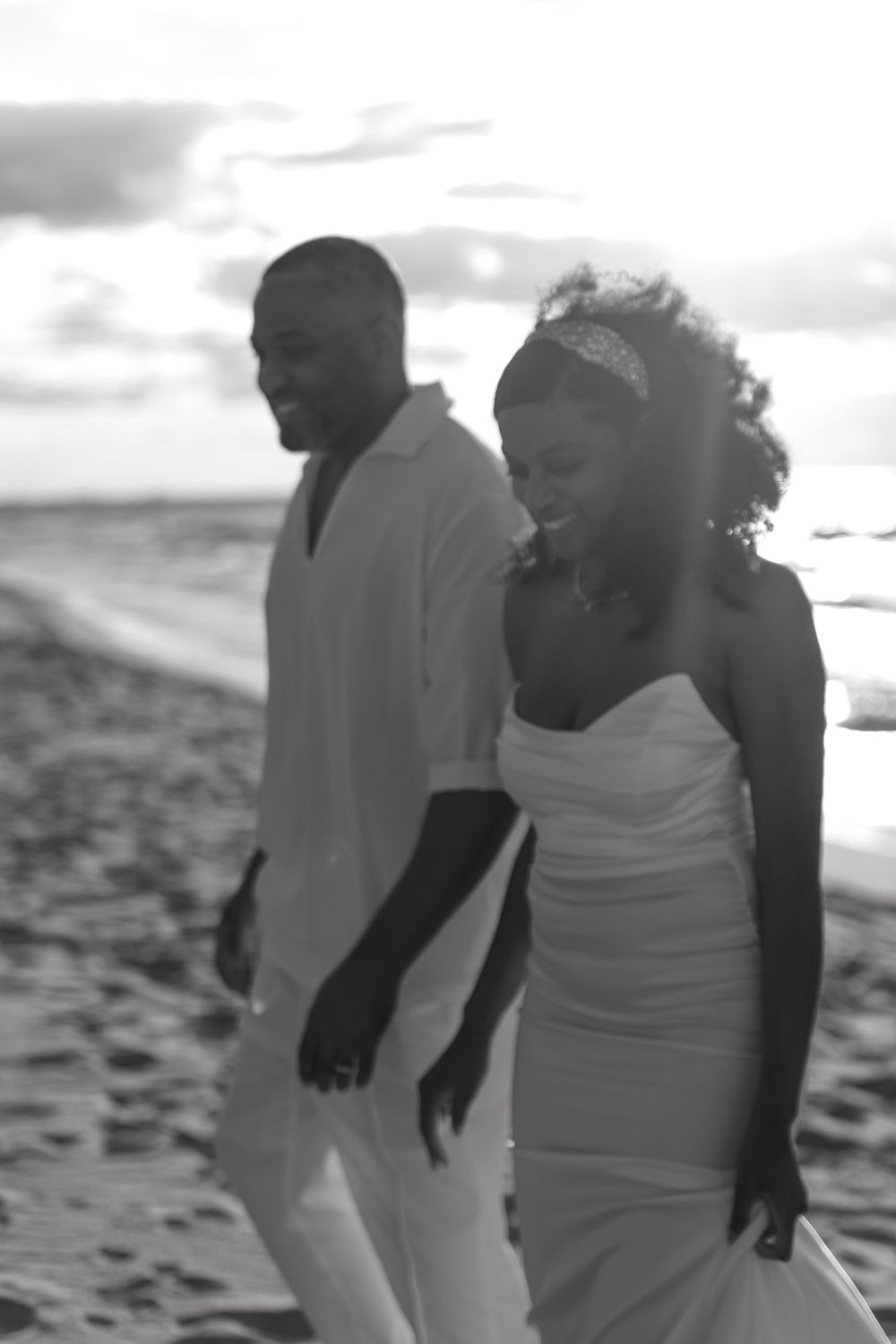 A couple walks hand in hand along a beach at sunset in black and white photography.