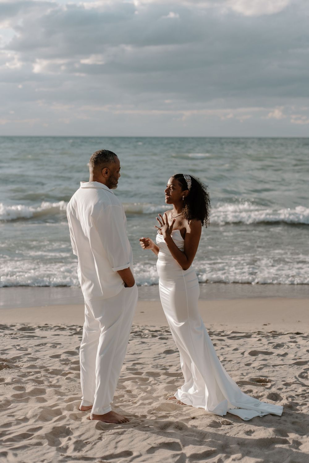 A couple in white outfits shares a romantic moment together on a sandy beach with ocean waves in the background.
