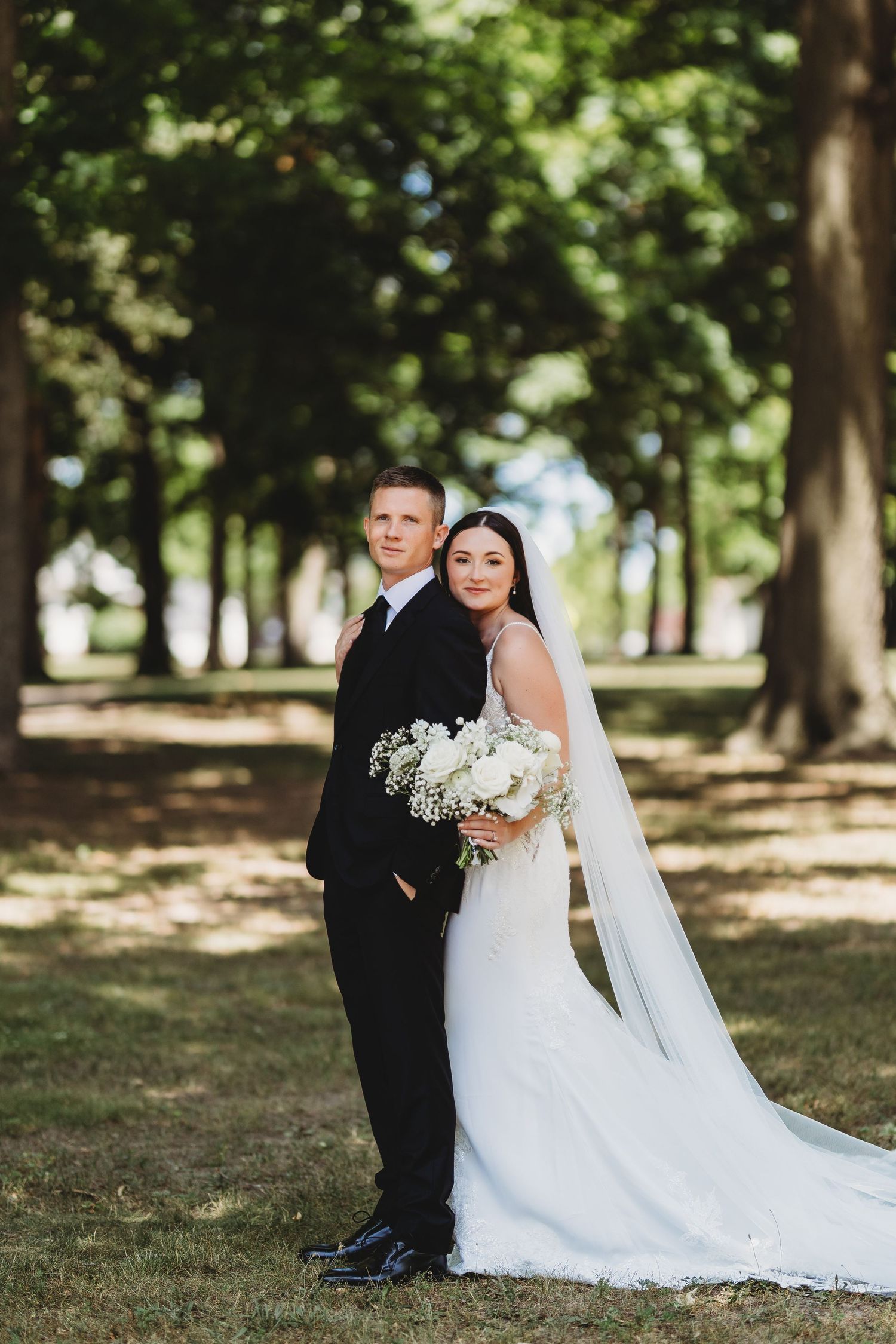 A romantic wedding portrait of a couple in a wooded park setting with the bride in a flowing white gown and veil.