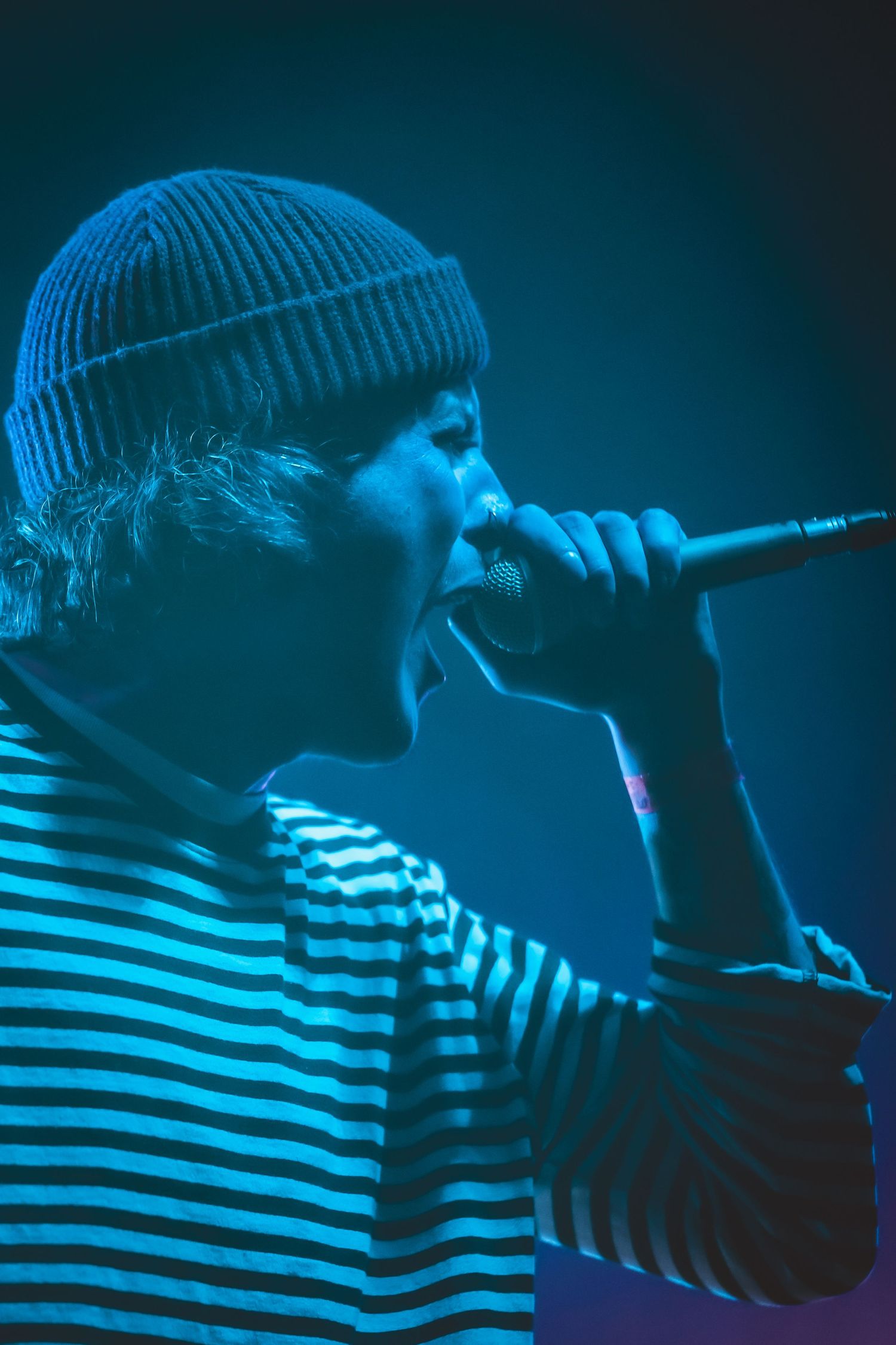 Performer in striped shirt and beanie hat singing on stage under dramatic blue lighting during live concert performance.