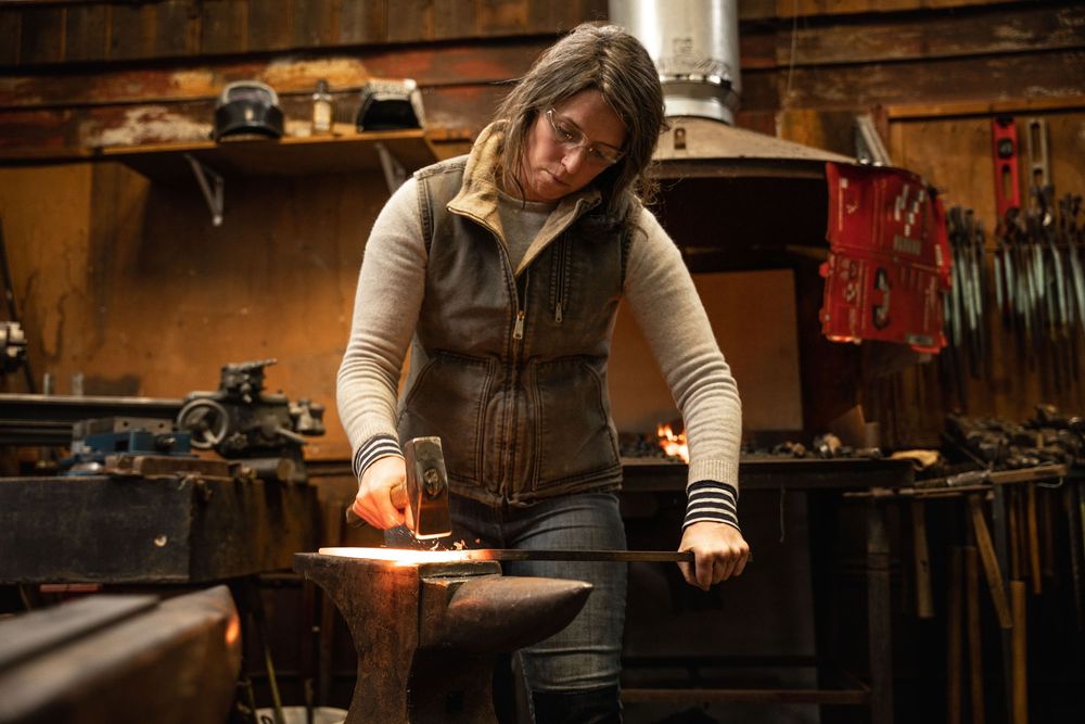 A woman working at an anvil in a rustic, tool-filled workshop; authentic brand photography for local artisans and makers in Tuscany.