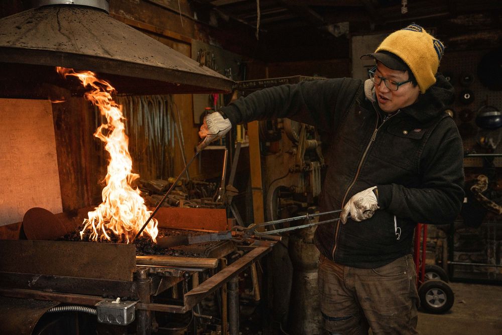 Blacksmith working with tongs near a forge fire in a workshop, wearing safety glasses and yellow hat.