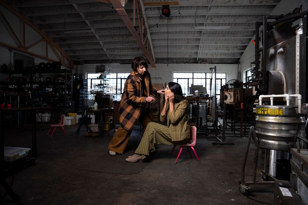 Two people pose in an industrial workshop setting, one standing in plaid, one seated on a red stool in olive green.