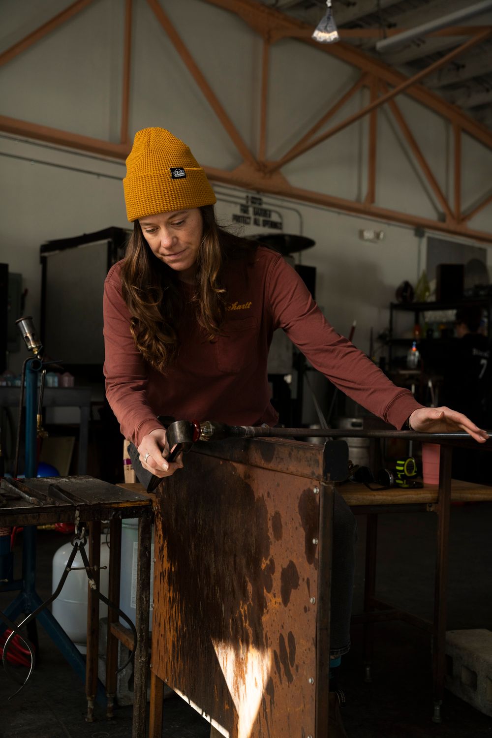 A female artisan in a yellow beanie and brown apron working in a Florence glass-blowing or craft studio; high-end artisan brand photography.