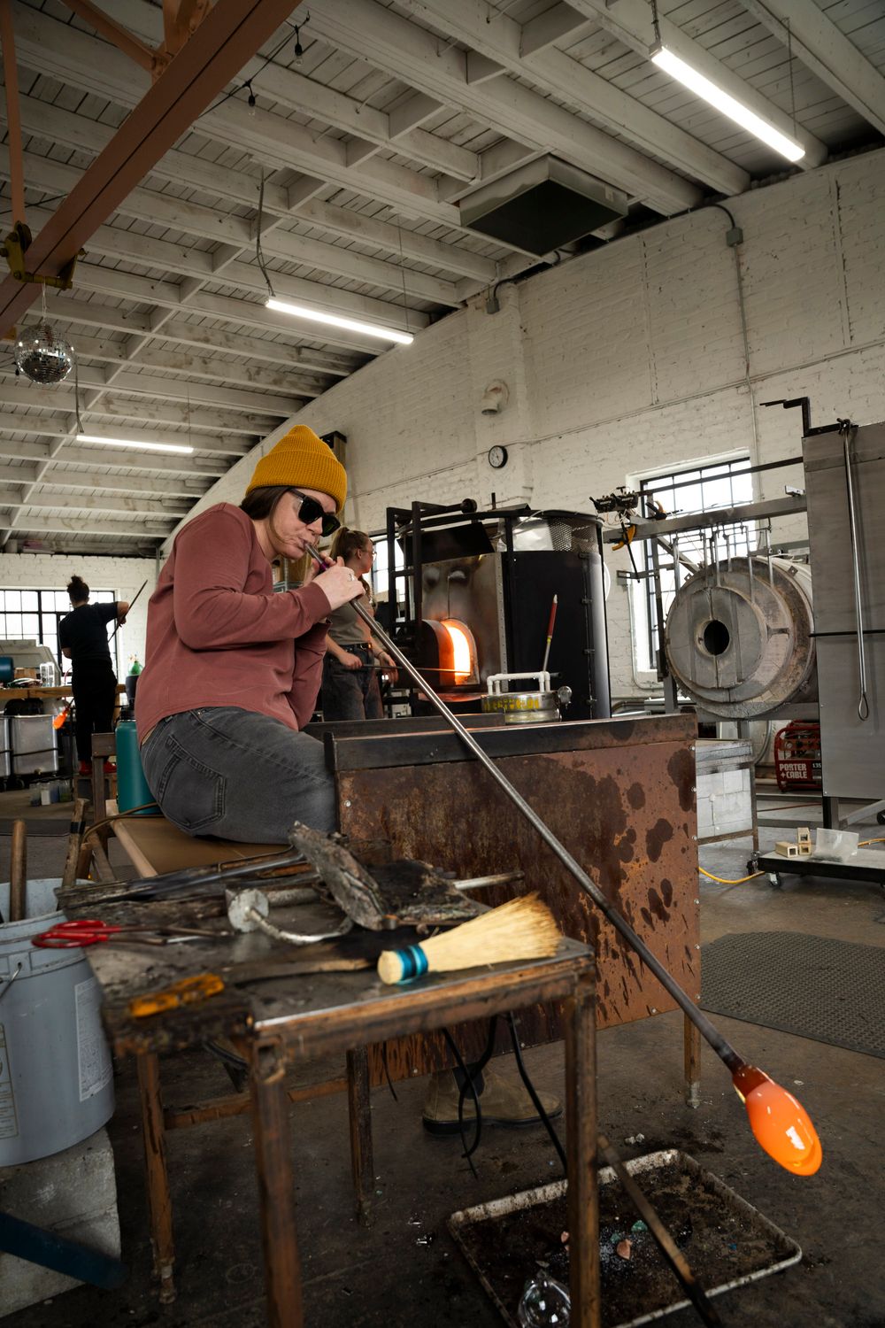 A glassblower in a yellow beanie shapes molten orange glass on a blowpipe at a workbench in an industrial studio.