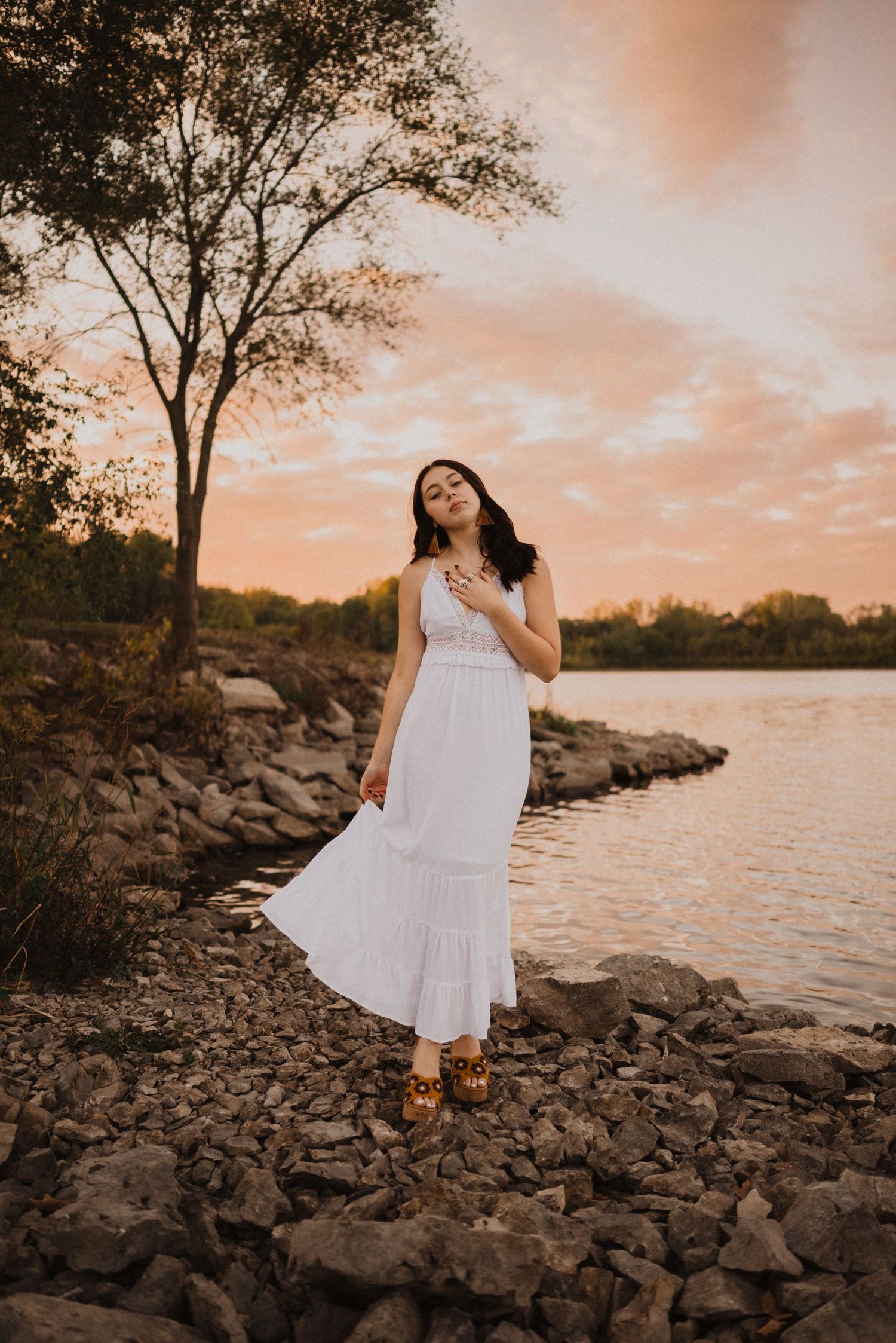 White dress figure stands on rocky shoreline at sunset with orange sky reflecting on calm waters.