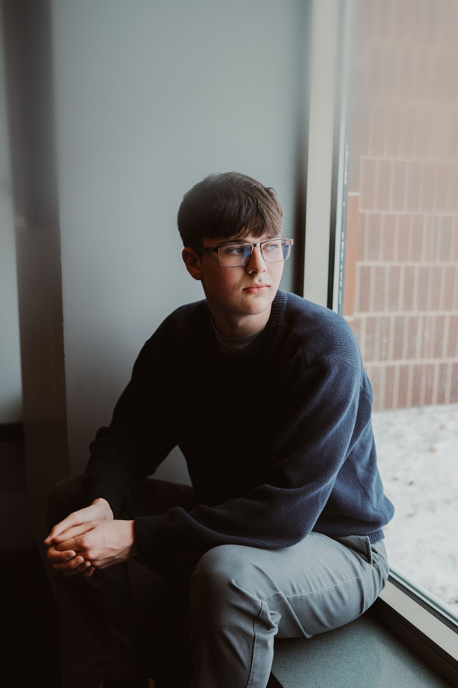 Person in dark blue hoodie sits pensively by window in moody lighting sequence.