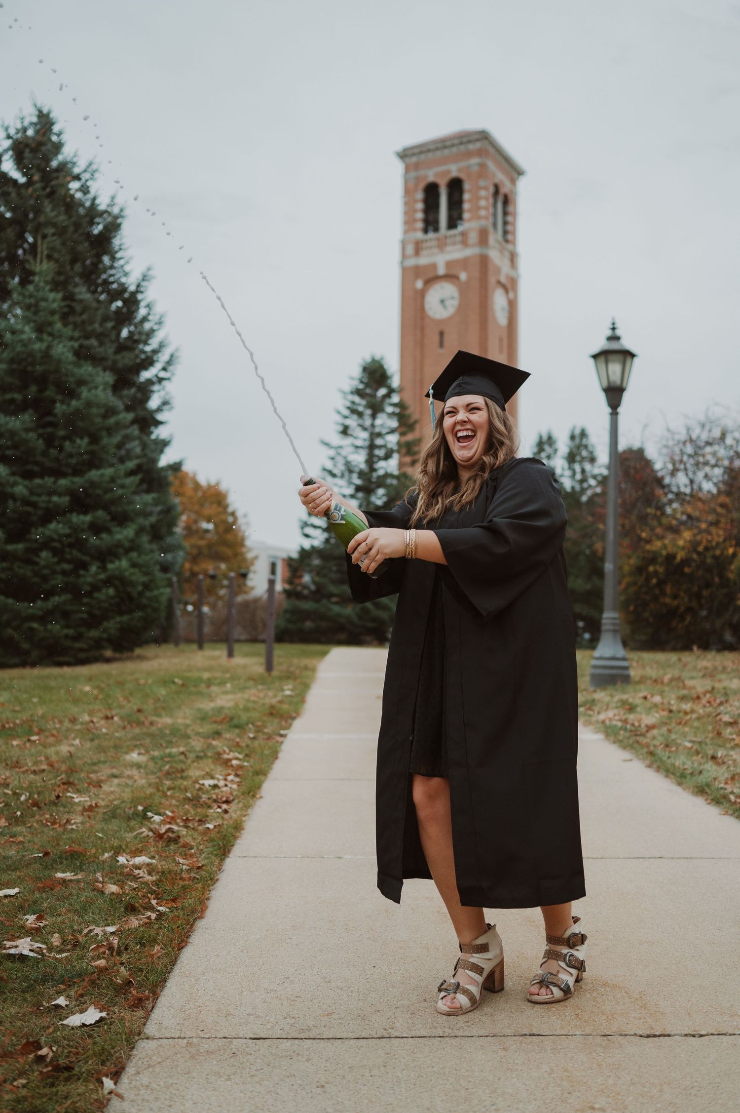 Graduate in black cap and gown poses playfully with wand near campus clock tower.