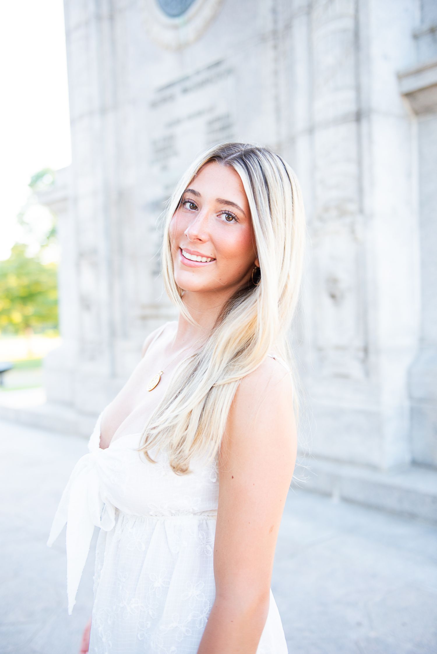 A young woman with long blonde hair smiles brightly while wearing a white dress against a light architectural backdrop.