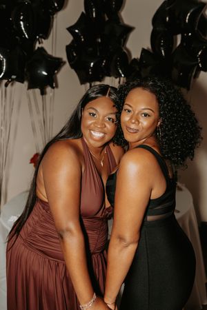 Two friends in brown and black dresses share joyful moments at an event with black balloons in the background.