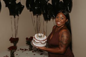 Guests celebrating at a party with decorative plants and a white cake visible in the background.