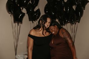 Two friends share a joyful moment in front of black balloons wearing black and brown dresses at an event.
