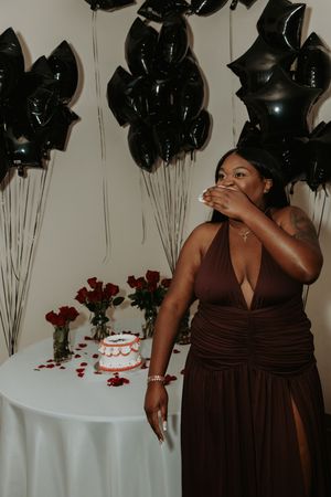 Woman in brown bridesmaid dress poses elegantly against wall with black balloons and white table setting.