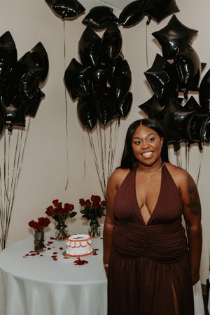 A person in a brown formal dress poses near a table with red roses and a cake at an elegant event.