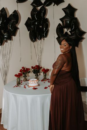 Bridesmaid in chocolate brown dress stands near white table with cake and red roses.