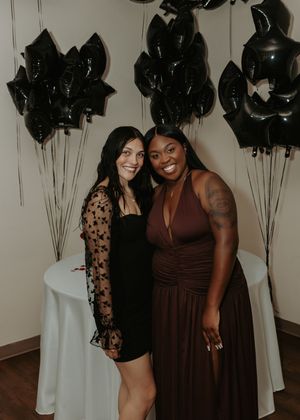 Two people share a joyful moment in front of black balloons wearing contrasting black and brown formal attire.