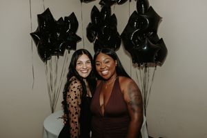 Two friends celebrate at a party with black balloons and decorative elements in the background.