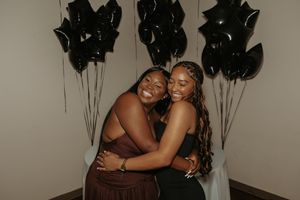 Two people embracing and smiling during a celebration with black balloons in the background.