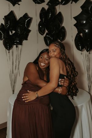 Friends embrace and laugh together at a celebration decorated with black balloons and white backdrops.
