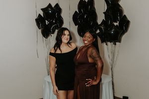 Two women pose together against a wall decorated with black balloons, wearing elegant cocktail dresses.