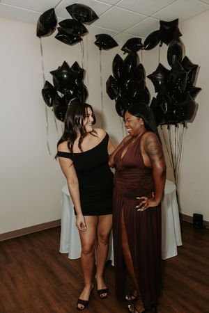 Two people in elegant dresses share candid moments against a backdrop of black balloons and white curtains.