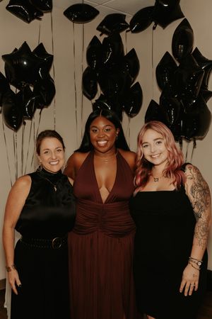 Three friends pose together at a party with black balloons decorating the background.
