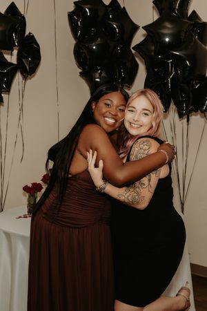 Two friends embrace and laugh together at celebration with black balloon backdrop.