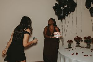 Friends gathered at a celebration with black balloon decor and rose petals visible in the scene.