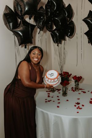 A celebratory table setup with black balloons, red rose petals, and a white birthday cake on a white tablecloth.