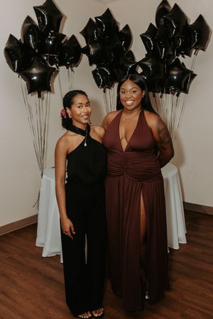 Two people pose together at a formal event with black balloons and wooden flooring.