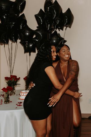 Friends laugh and embrace at a celebration with black balloons and red roses in an elegant indoor setting.