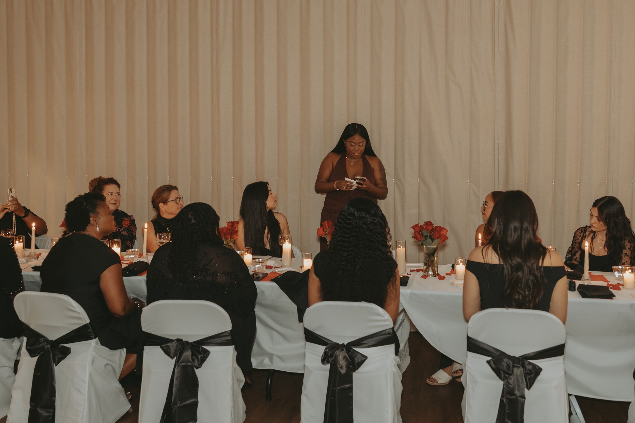 A group gathers around an elegant white-clothed table with black chair sashes at a formal dining event.