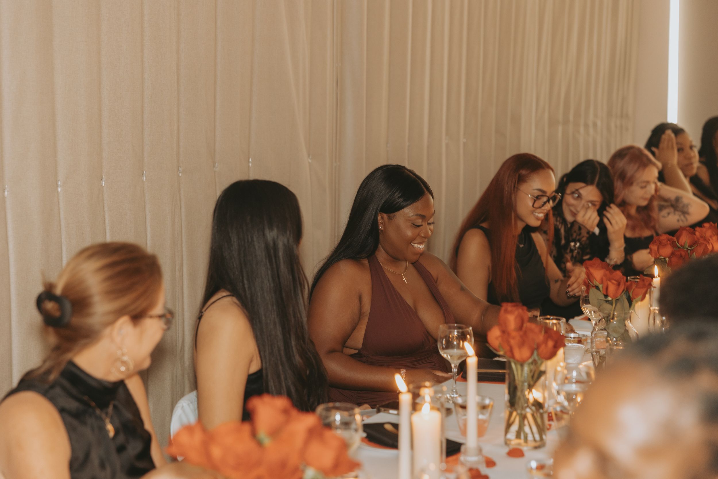 Wedding guests laugh and celebrate together at a long reception table decorated with white linens and floral centerpieces.