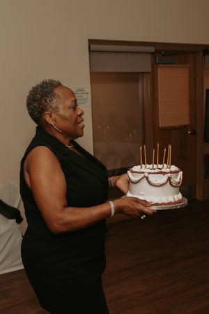 Someone in black holds a decorated birthday cake with lit candles in a dimly lit room.