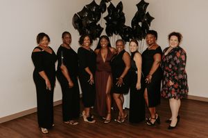 A group of eight people pose together wearing elegant black dresses and one in brown against a white wall with dark decor.