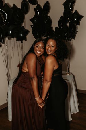 Two friends in formal attire share a joyful moment at an event with black star decorations in the background.