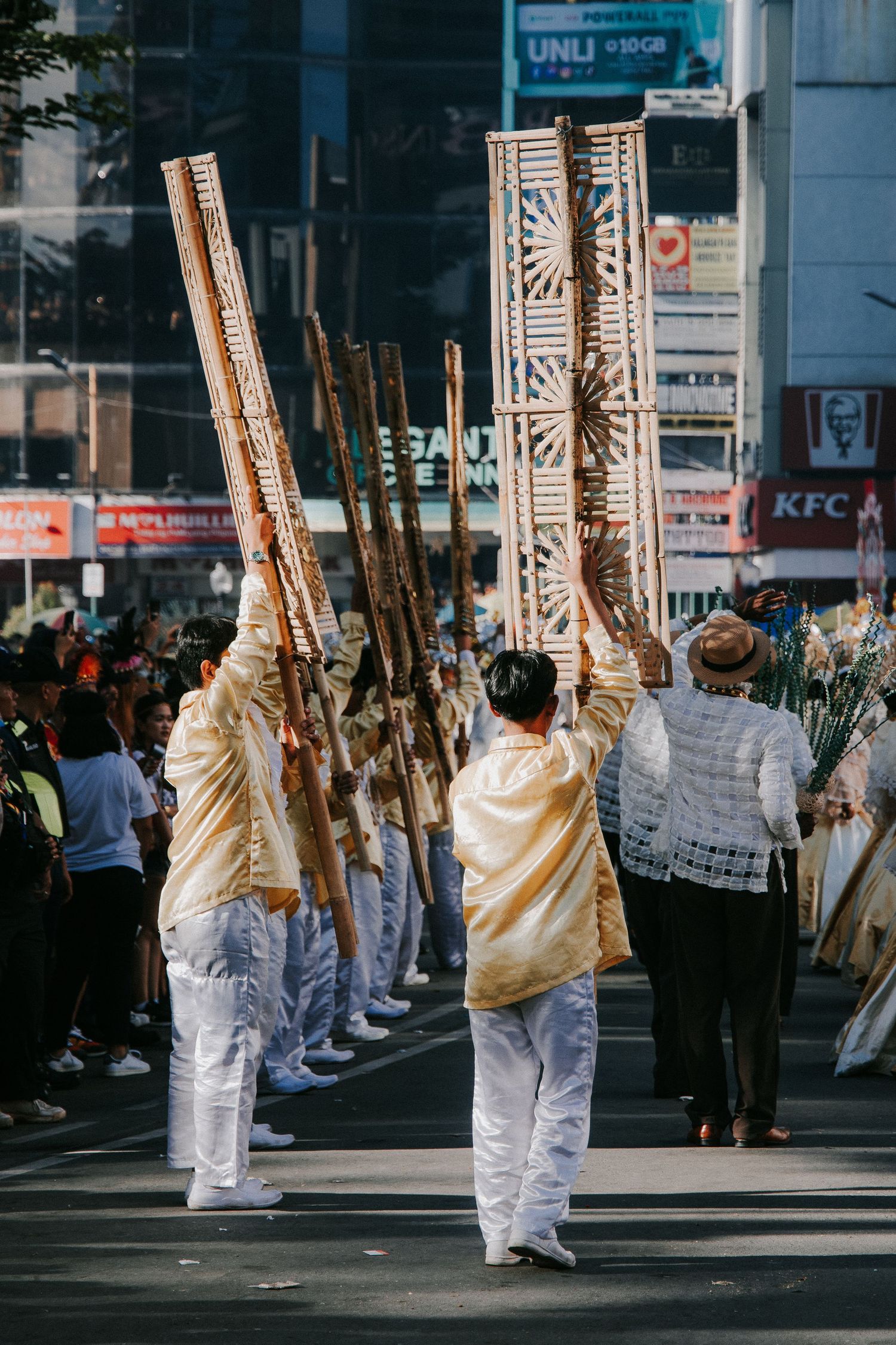 People in white clothing carrying tall ceremonial poles during a street procession.
