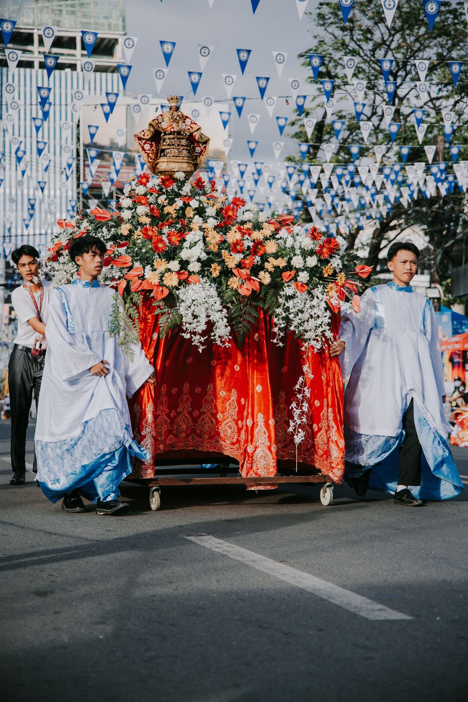Festival performers in white robes dance in front of red and floral decorations under blue banners.
