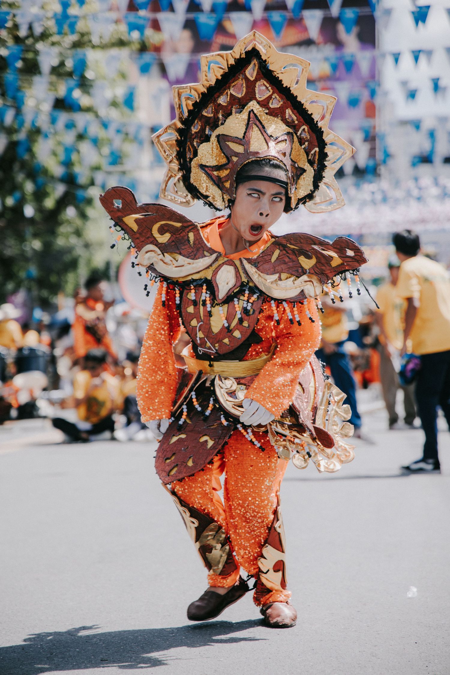 Performer in orange costume and elaborate headdress dances energetically during street festival.