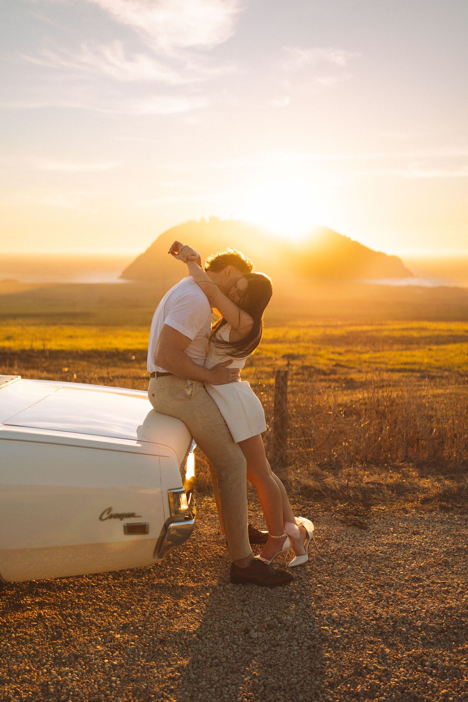 Sunset engagement session with couple by boat against mountain backdrop.