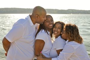 Happy Black family of four hugging and smiling together on a sunny beach, all wearing white shirts.