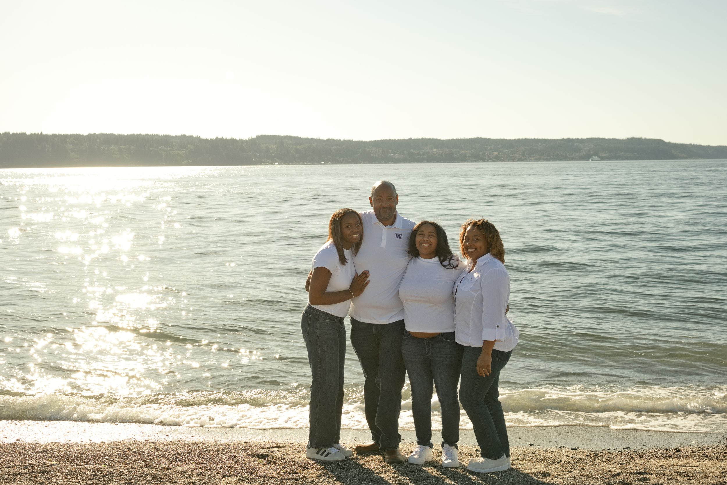 Four people smiling together on a sunny beach with calm water and hills in the background.