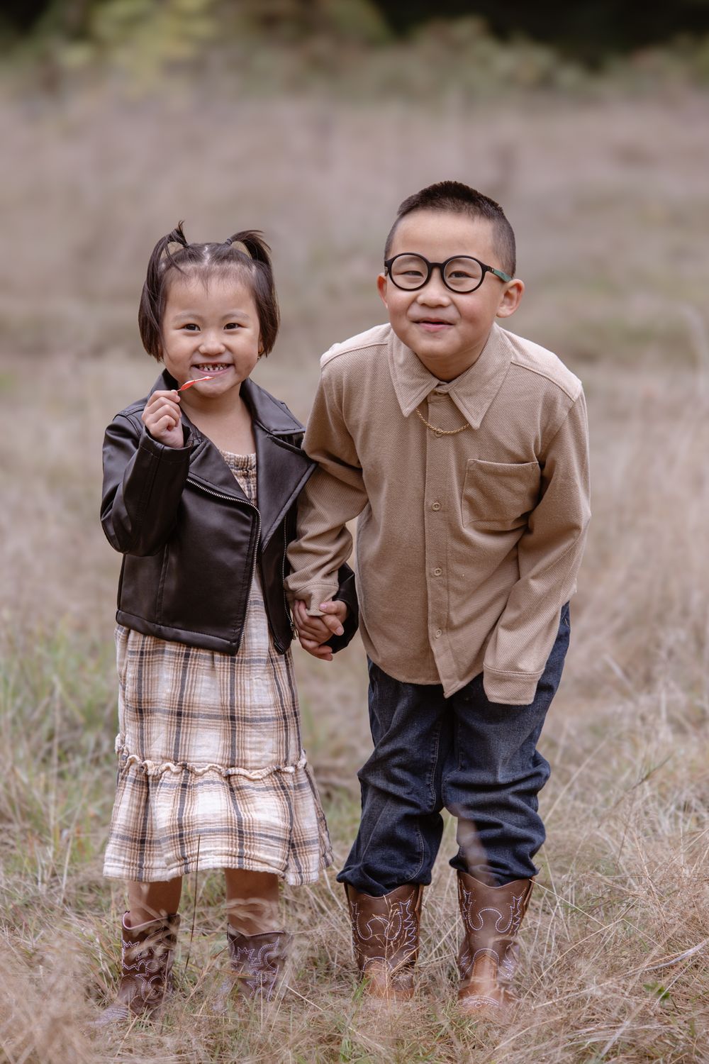 Two young children in matching brown and plaid outfits stand together in a grassy field.