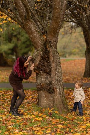 A playful game of peek-a-boo around a large tree trunk surrounded by fallen autumn leaves.