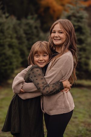 Two young girls in coordinated outfits share warm embraces while posing outdoors against a natural backdrop.