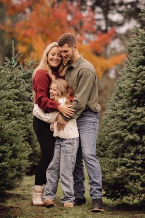 A family shares a tender moment among evergreen trees with vibrant autumn foliage in the background.
